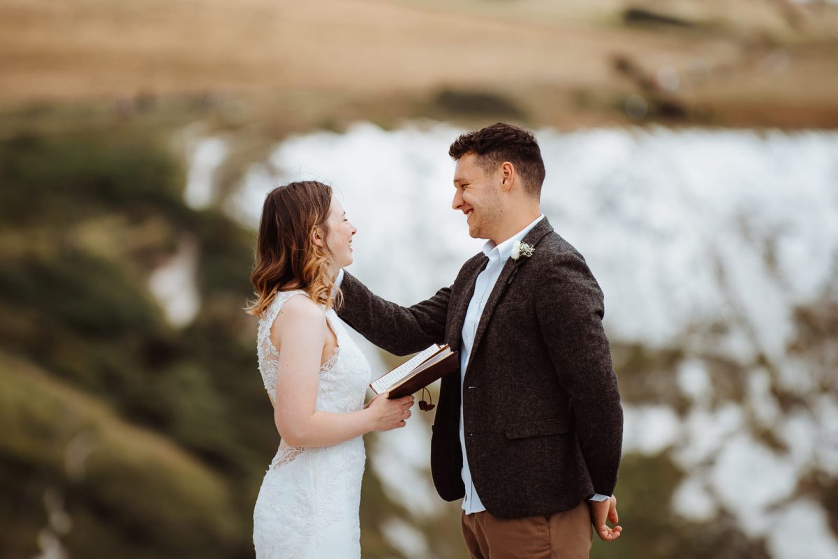 A couple sharing their vows on a cliff. The bride id reading her love words, smiling and enjoying the moment. The groom is gently stroking her face