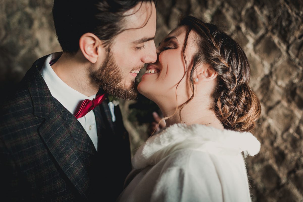 Close portrait of a couple smiling and kissing. The bride wears a winter white coat. The groom wears a British attire