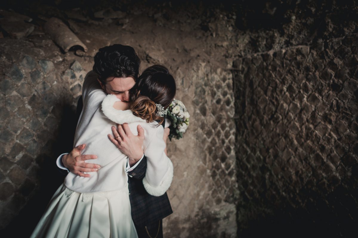 Couple hugging inside an ancient ruins in Rome. The bride is wearing a white winter bridal coat. The groom is holding her, with his face gently rubbing on her shoulder