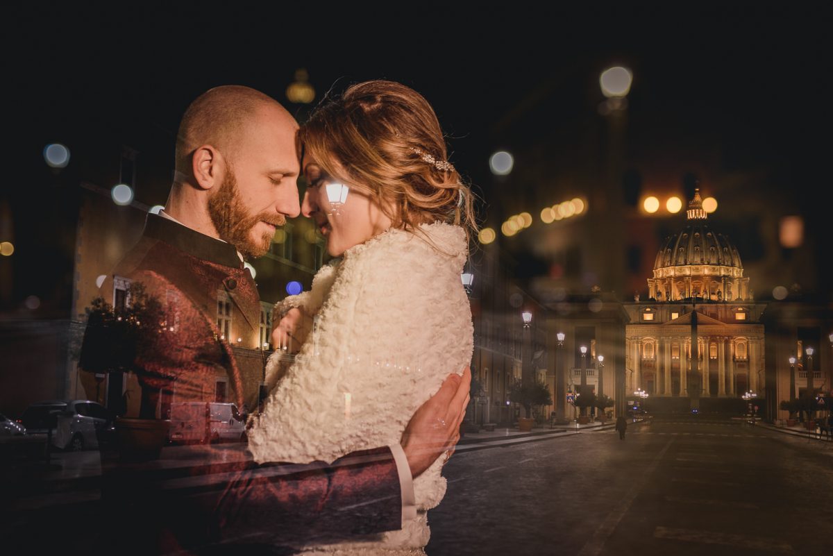 Couple that gently hug each other in front of San Pietro church in Rome at night. The bride is wearing a white wool cape, and the groom is wearing a red silk jacket. There are fairy lights spreading around them