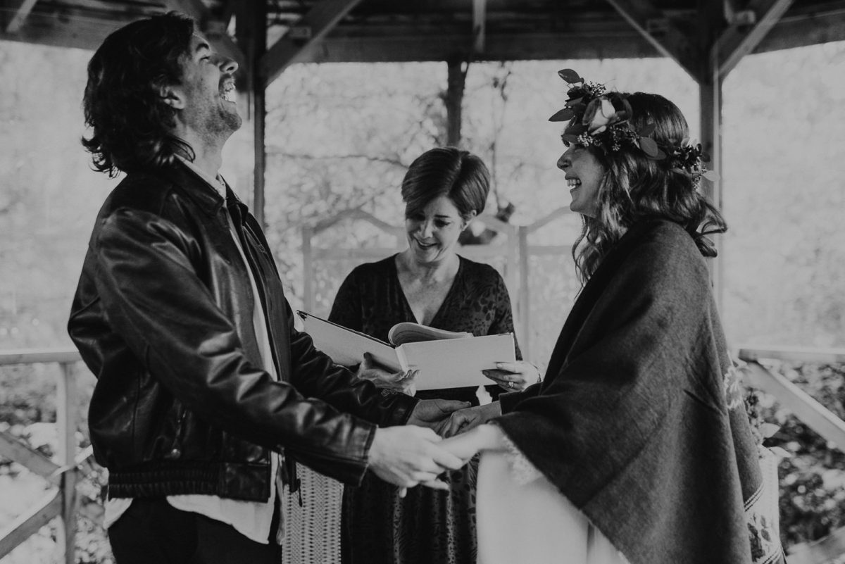 A couple getting married under a gazebo in Lake District. The bride has a flower crown, and a wool cloak. The groom wear a vintage leather jacket. They are holding hands, and laughing