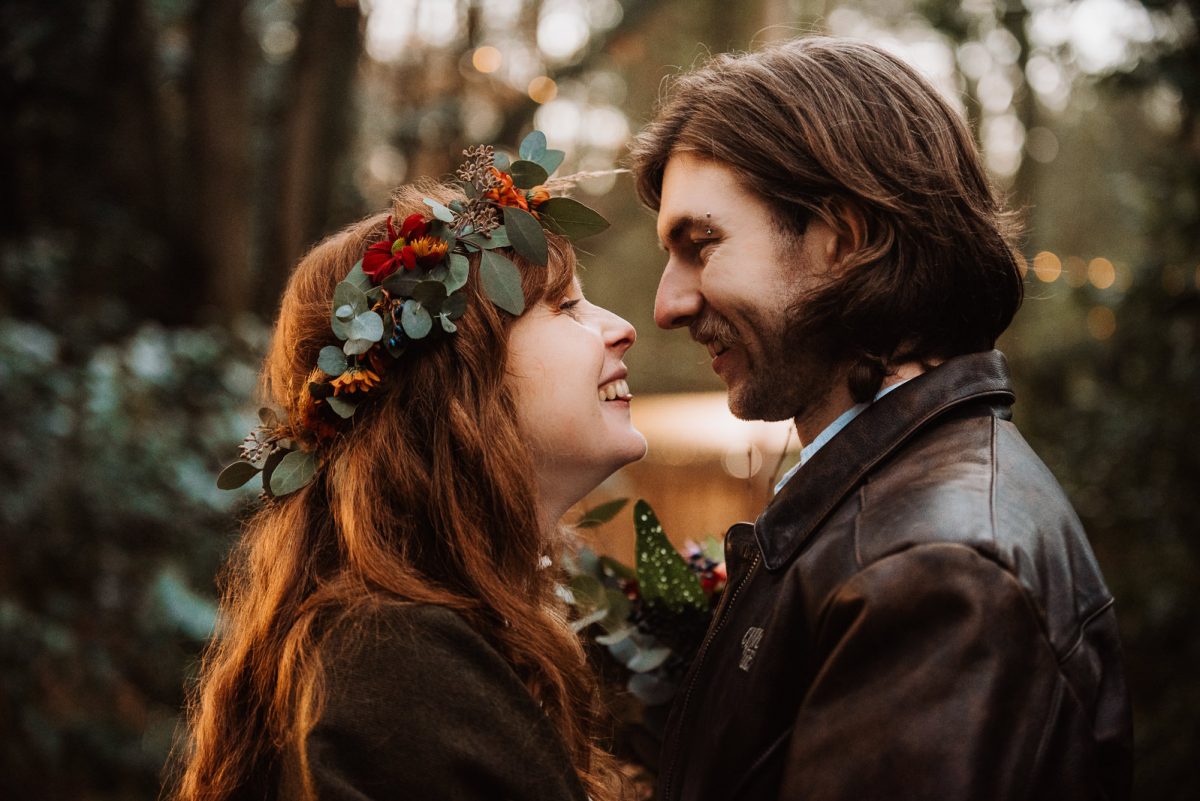Portrait of a just married couple. The bride wear a flower crown, and she is smiling and so happy. The groom wear a vintage leather jacket, and has a eyebrows piercing